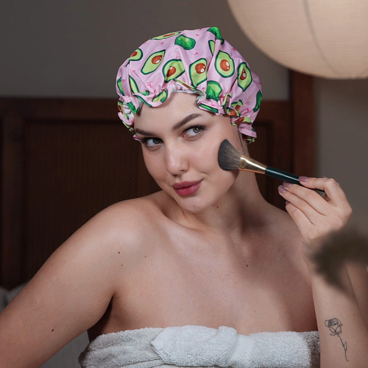 Woman applying makeup with a brush, wearing a SMUG avocado-patterned shower cap and towel.