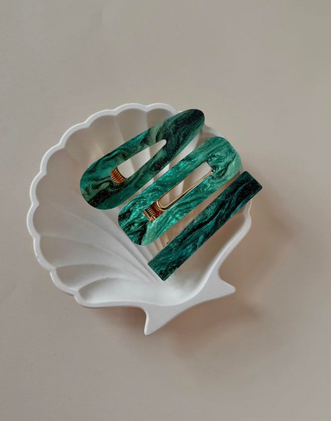 A set of three green hairclips on a white shell-shaped stand against a light background