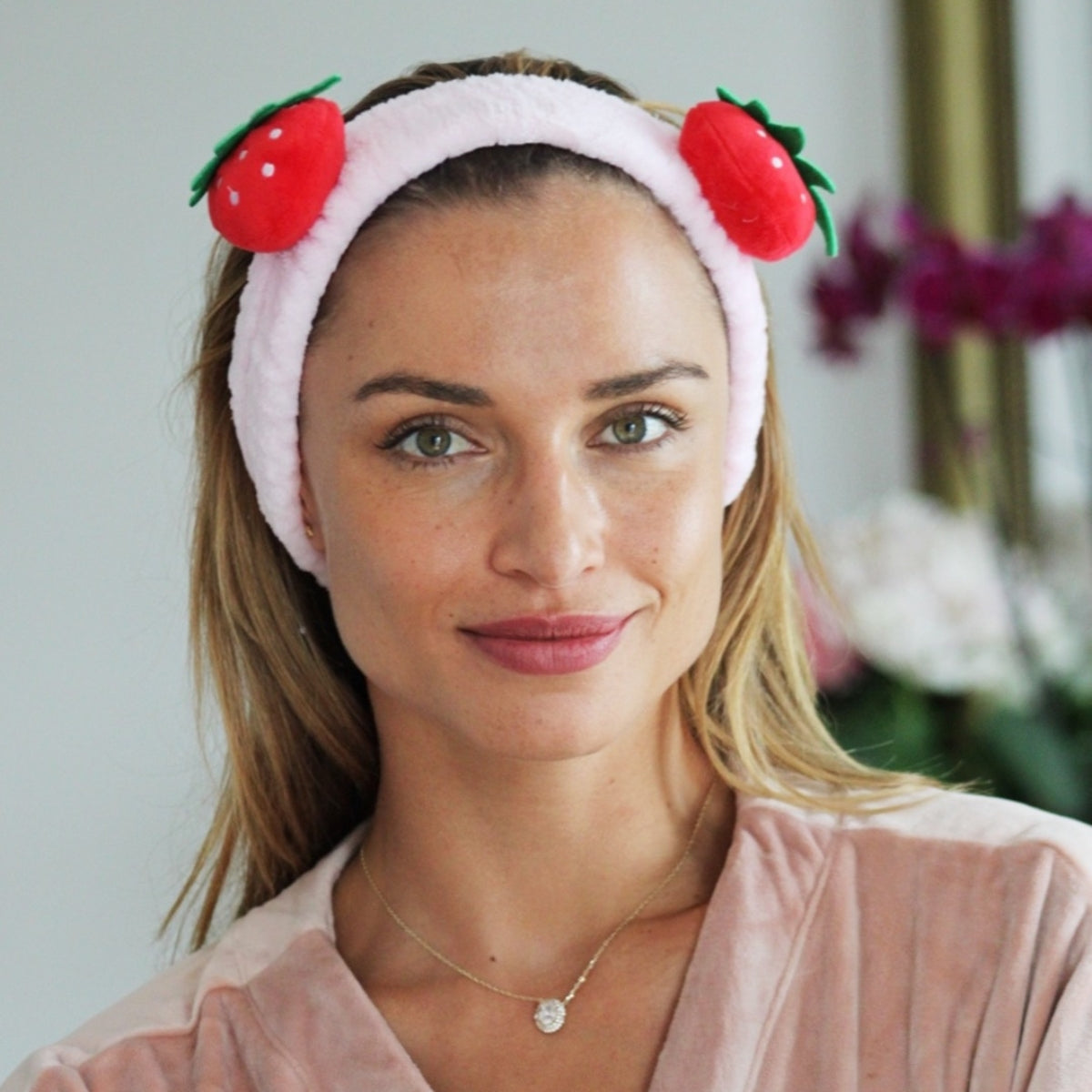 Woman wearing a pink plush SMUG strawberry themed spa headband against a neutral background