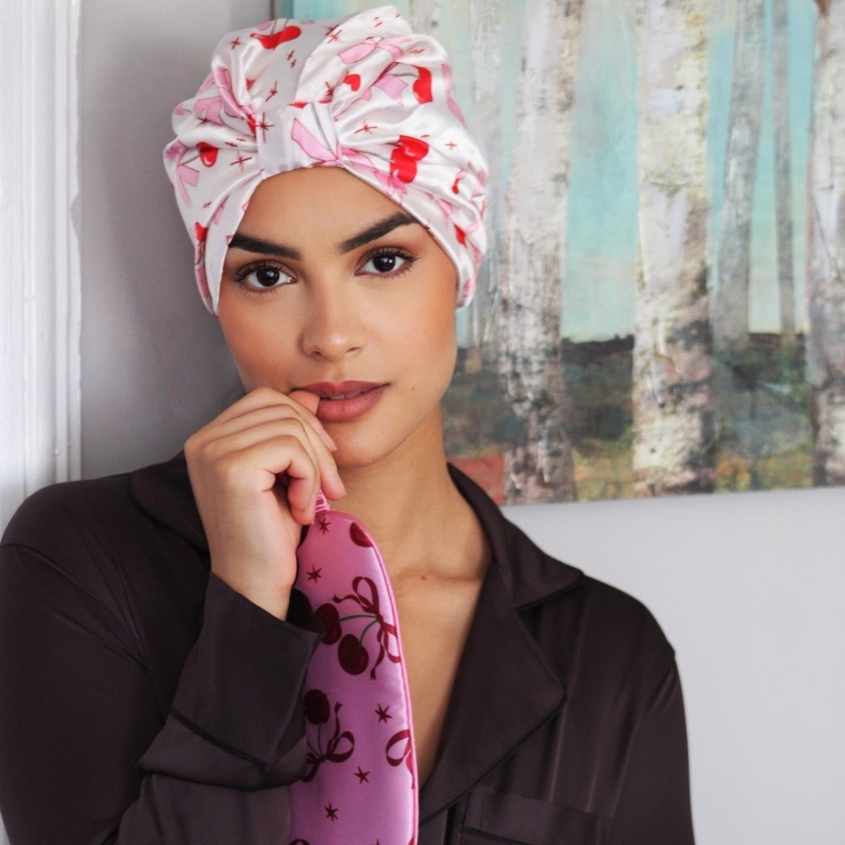 Model wearing a SMUG Cherry Bows satin sleep turban and holding a Pink Cherries sleep mask.
