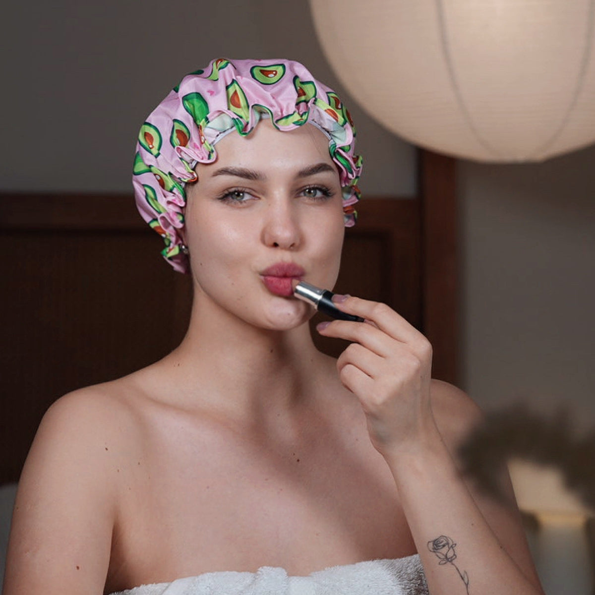 Woman applying lipstick with a colourful SMUG avocado print shower cap on her head.