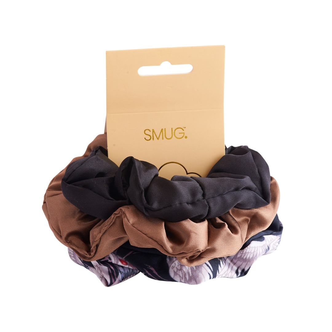 Set of three scrunchies in an elegant crane-printed design, sumptuous mocha satin and timeless black on a white background with 'SMUG' branding.