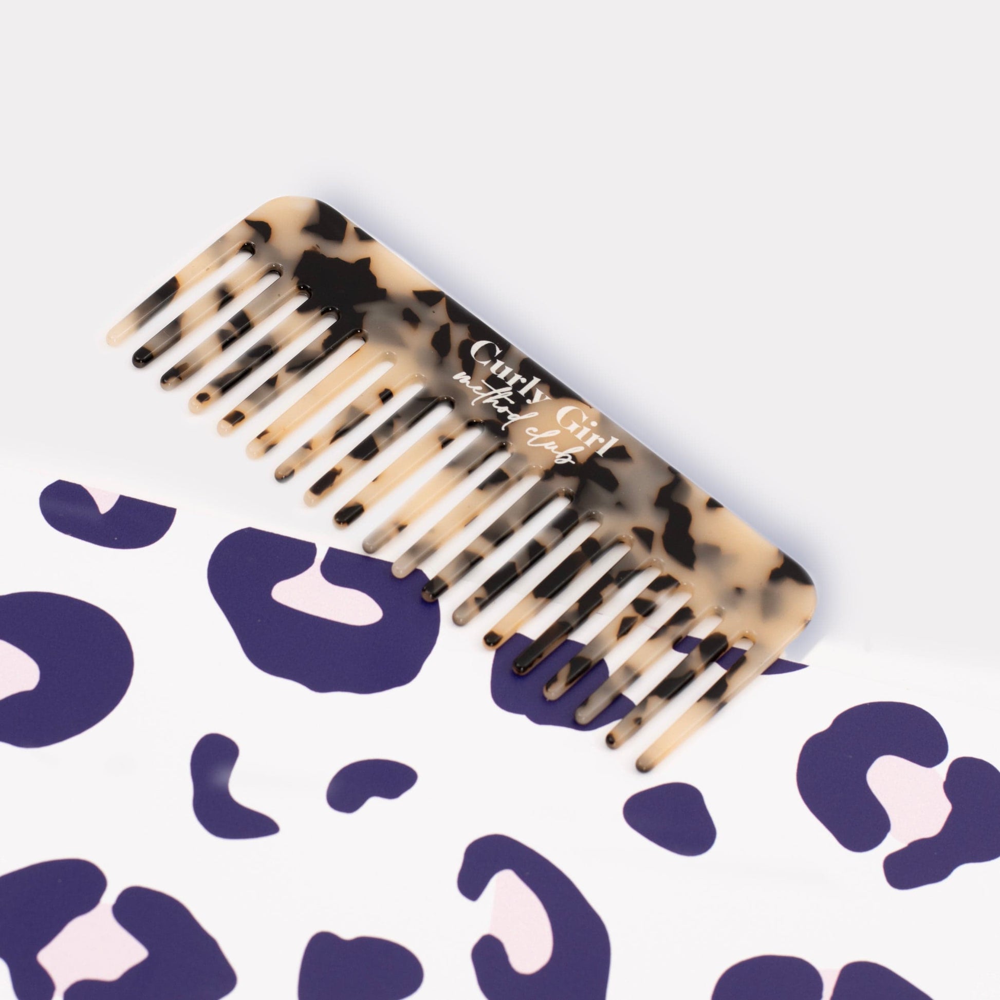 Tortoiseshell comb with 'Curly Girl Method Club' text on a white background with purple abstract patterns